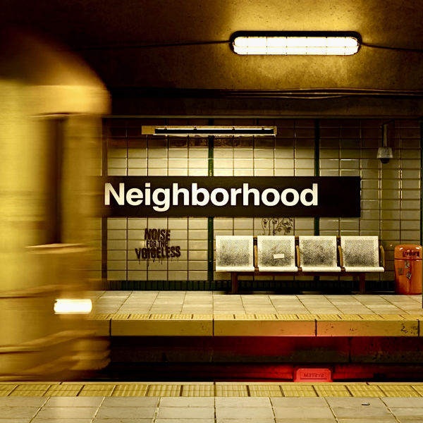 Cover image for the song 'Neighborhood' featuring a subway station with a blurred train passing by. The station sign reads 'Neighborhood' in bold white letters, and the band's name, 'Noise For The Voiceless,' is subtly visible on the tiled wall behind empty metal chairs.